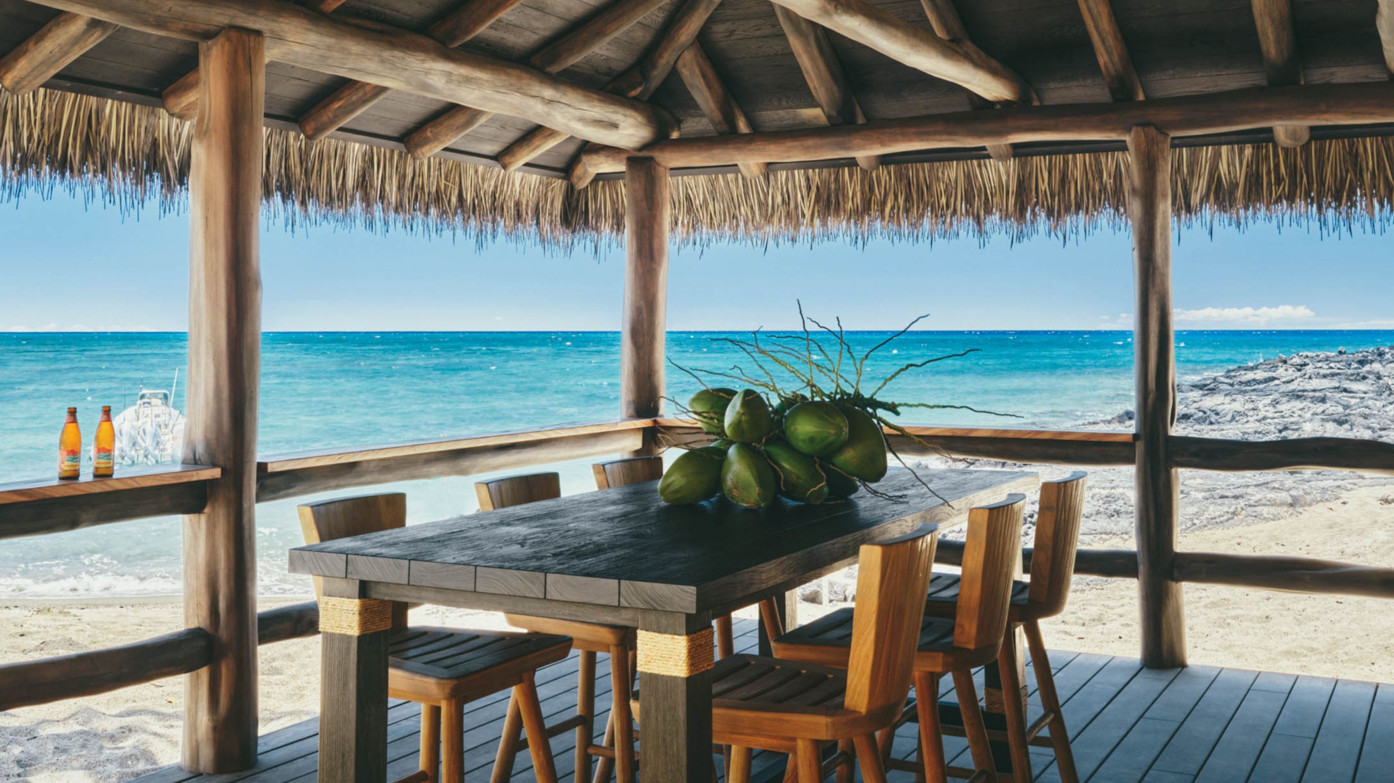 A beachside hut with a thatched roof, featuring a table with coconuts and drinks, overlooking the ocean.